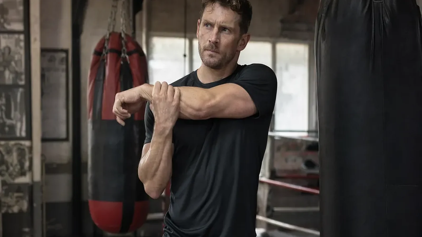 Accountant in dark training kit loosening tight shoulders beside heavy bags after desk work