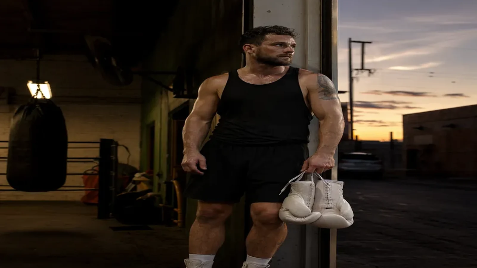 Boxer standing in a gym doorway after training holding white gloves at dusk