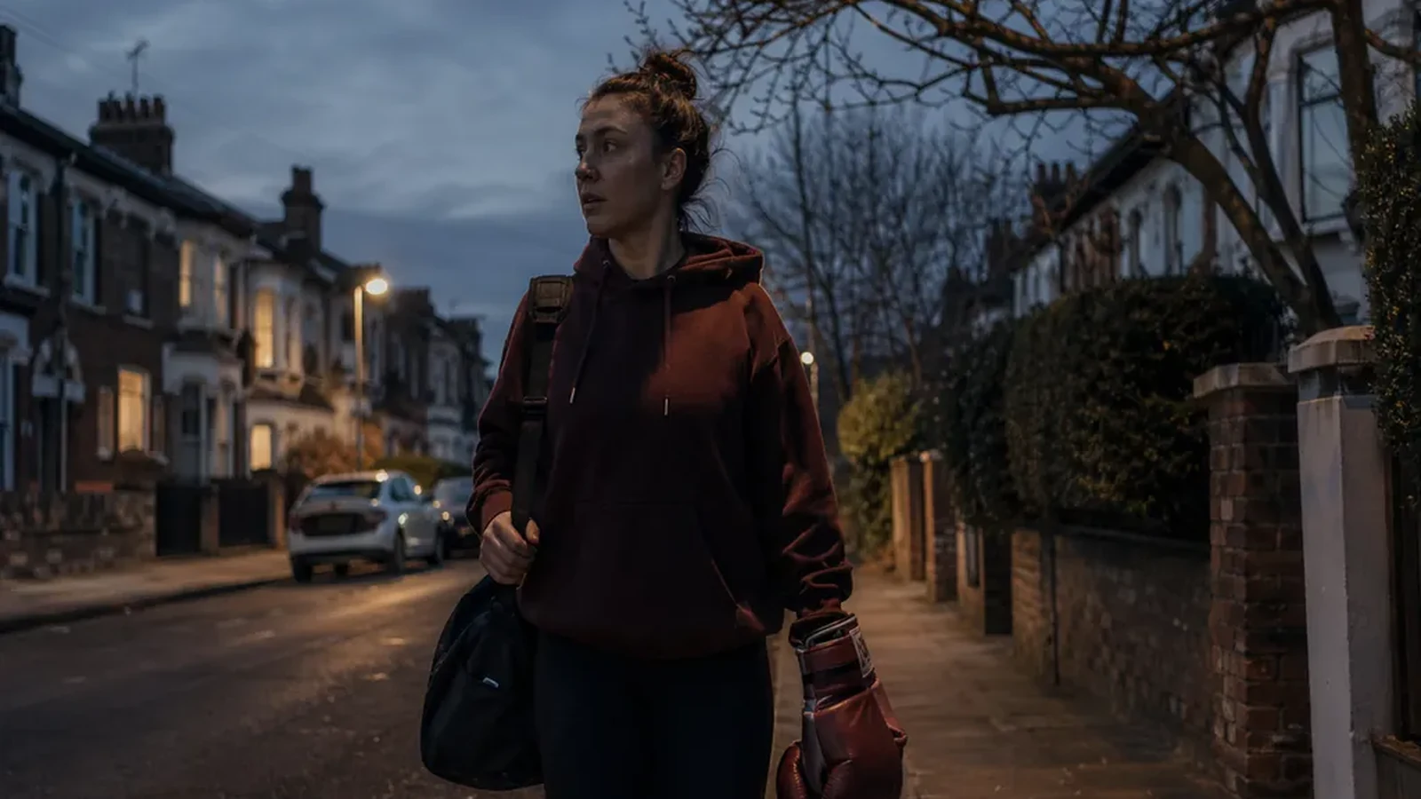 Woman walking home with a gym bag and boxing glove through a residential street at dusk