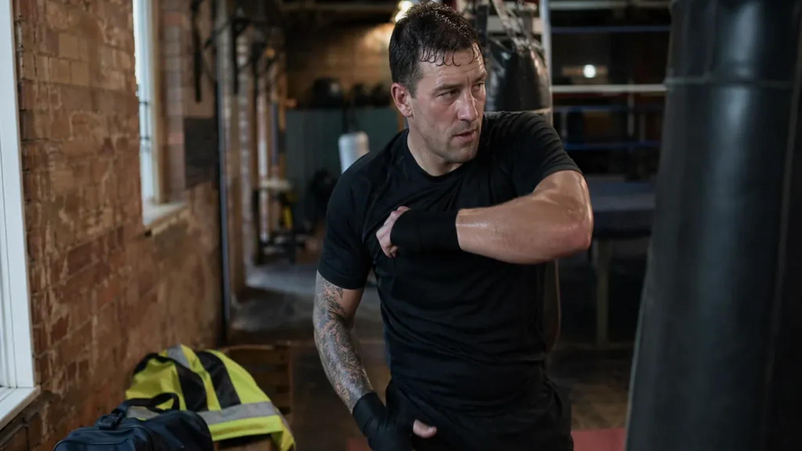 Emergency-service worker in dark training kit wrapping hands beside heavy bags before a boxing session