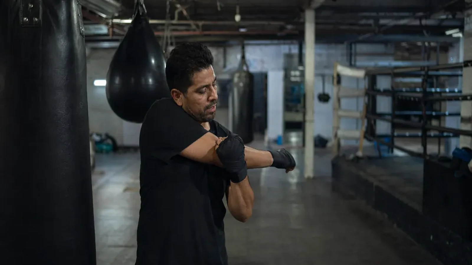 Man stretching tight shoulders beside heavy bags before evening boxing training