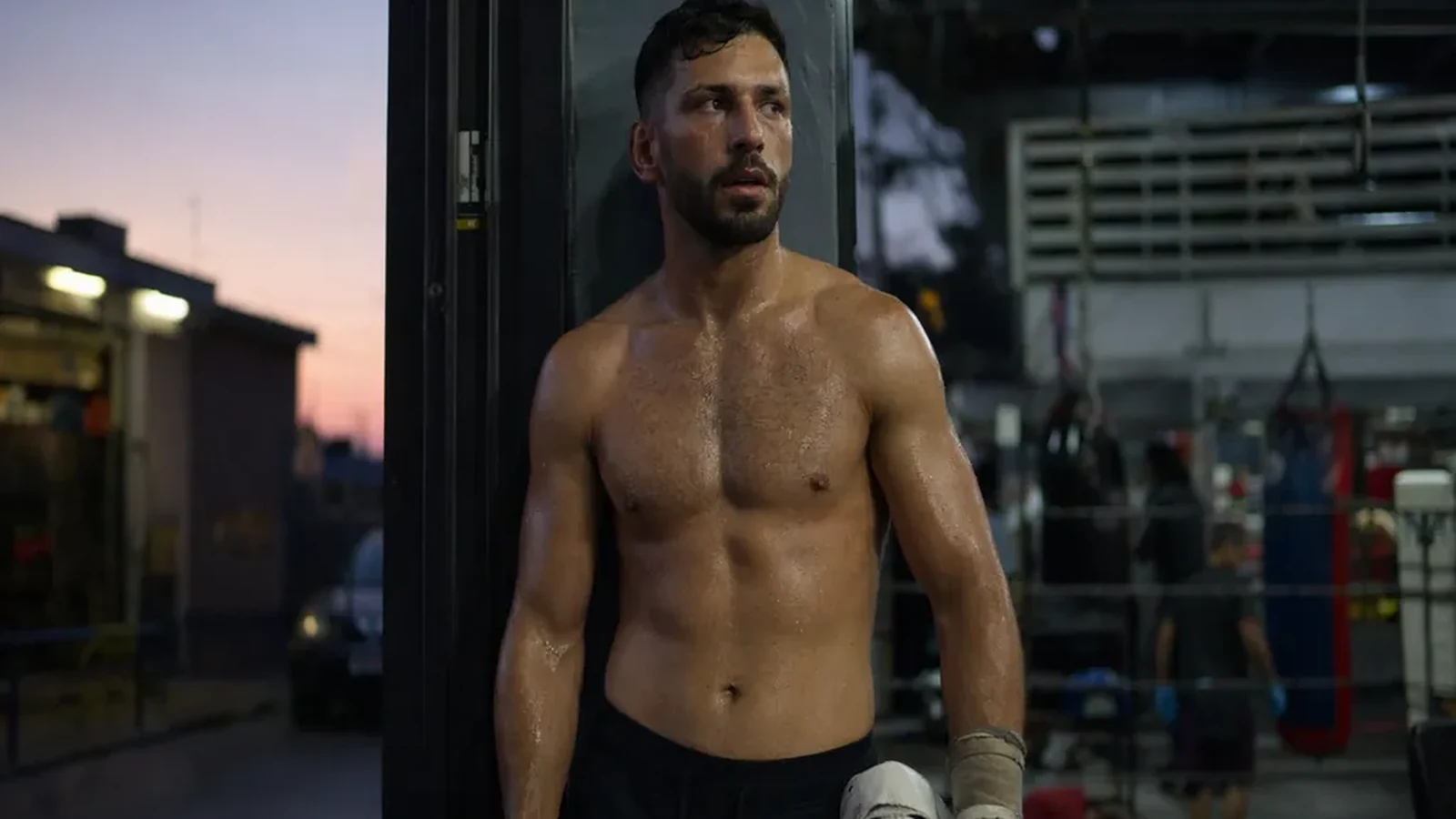 Boxer standing in a gym doorway after training holding white gloves at dusk
