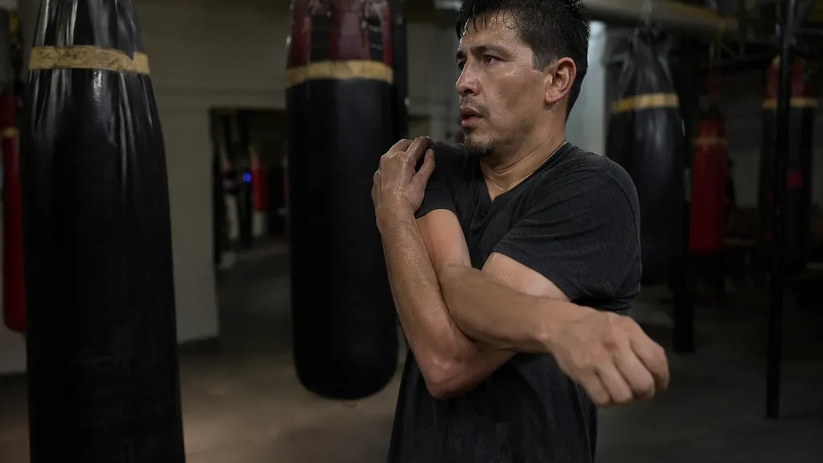 Man loosening tight shoulders before evening boxing training beside heavy bags