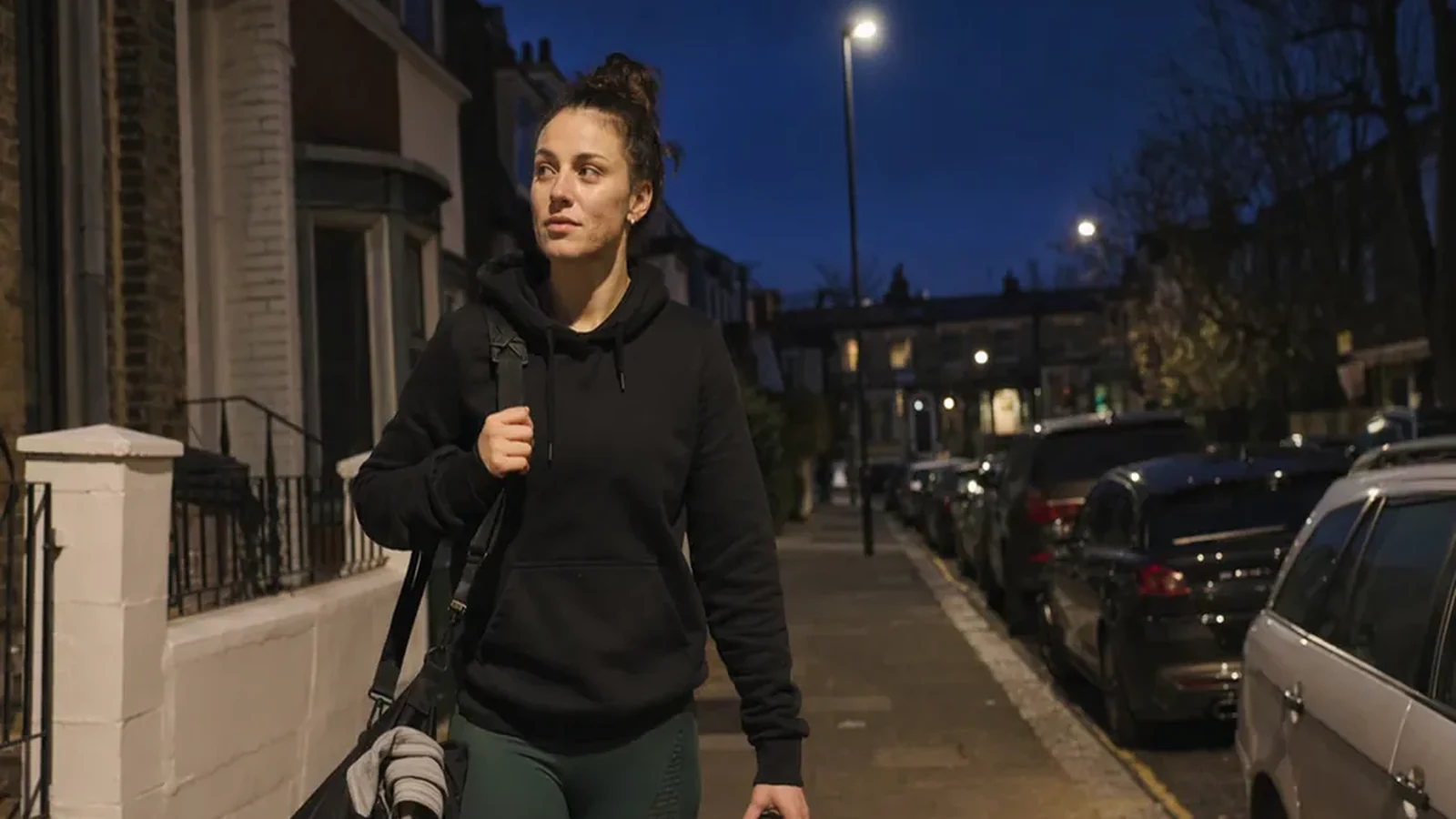 Woman walking home after evening training with a gym bag through a residential street at dusk