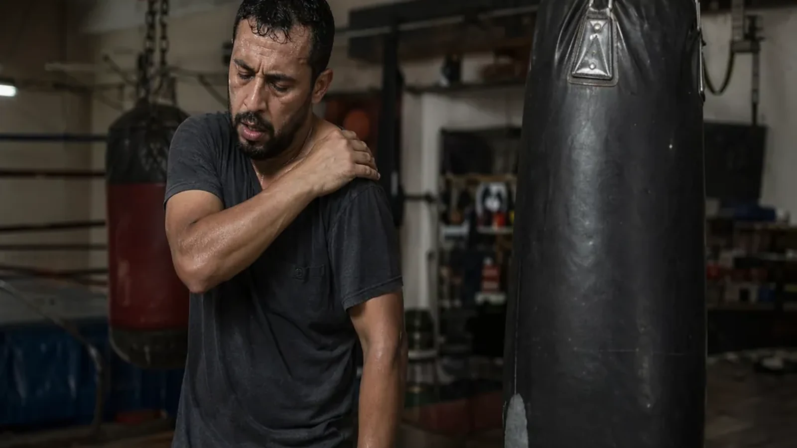 Man rubbing a sore shoulder beside heavy bags after boxing training