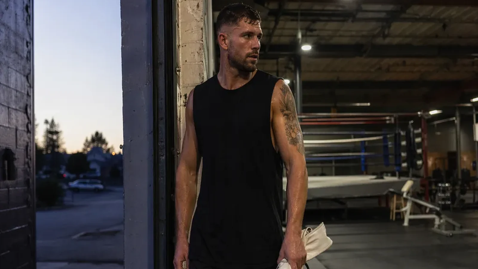 Boxer standing in a gym doorway after training holding white gloves at dusk