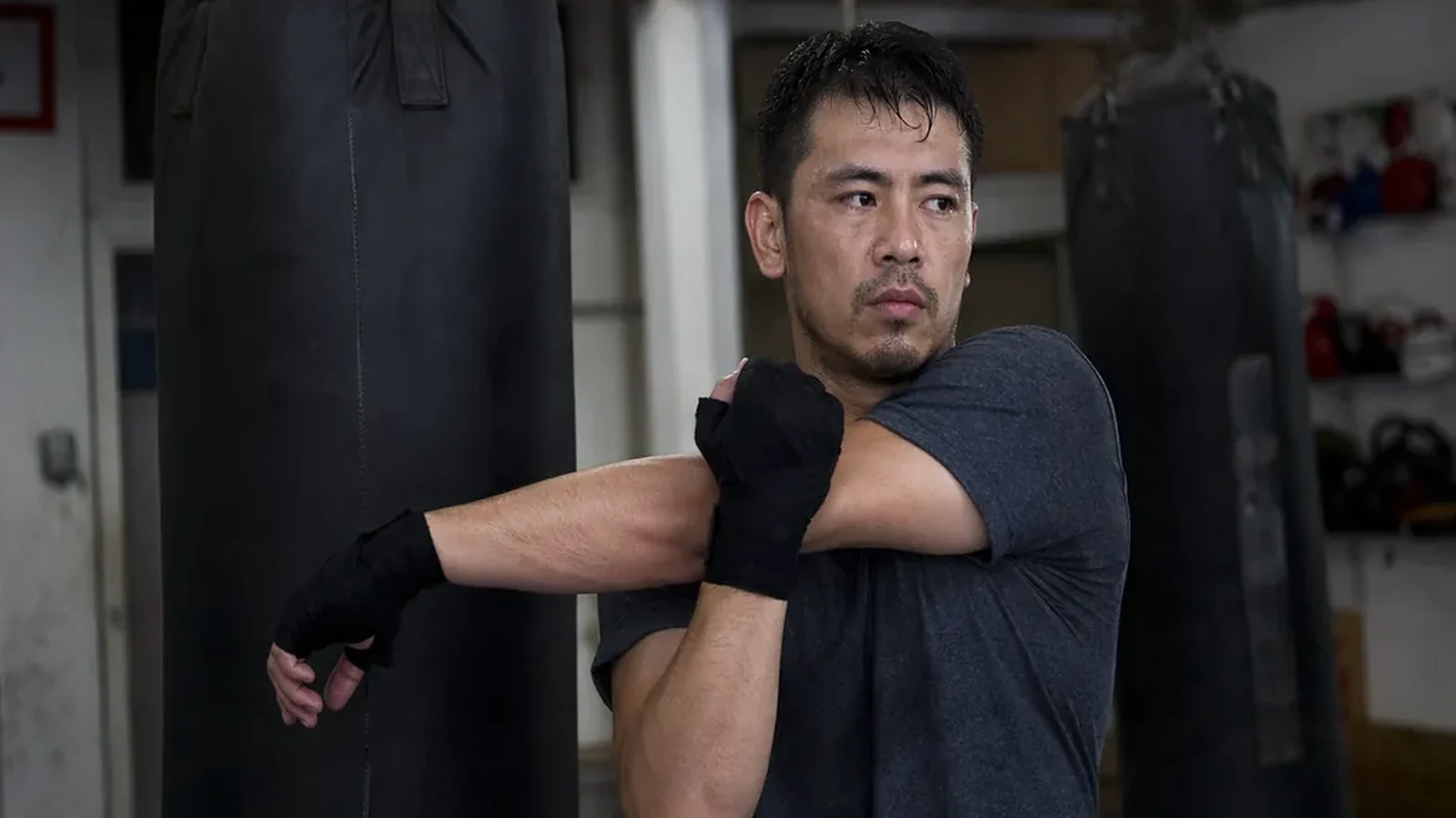 Man stretching tight shoulders beside heavy bags before evening boxing training