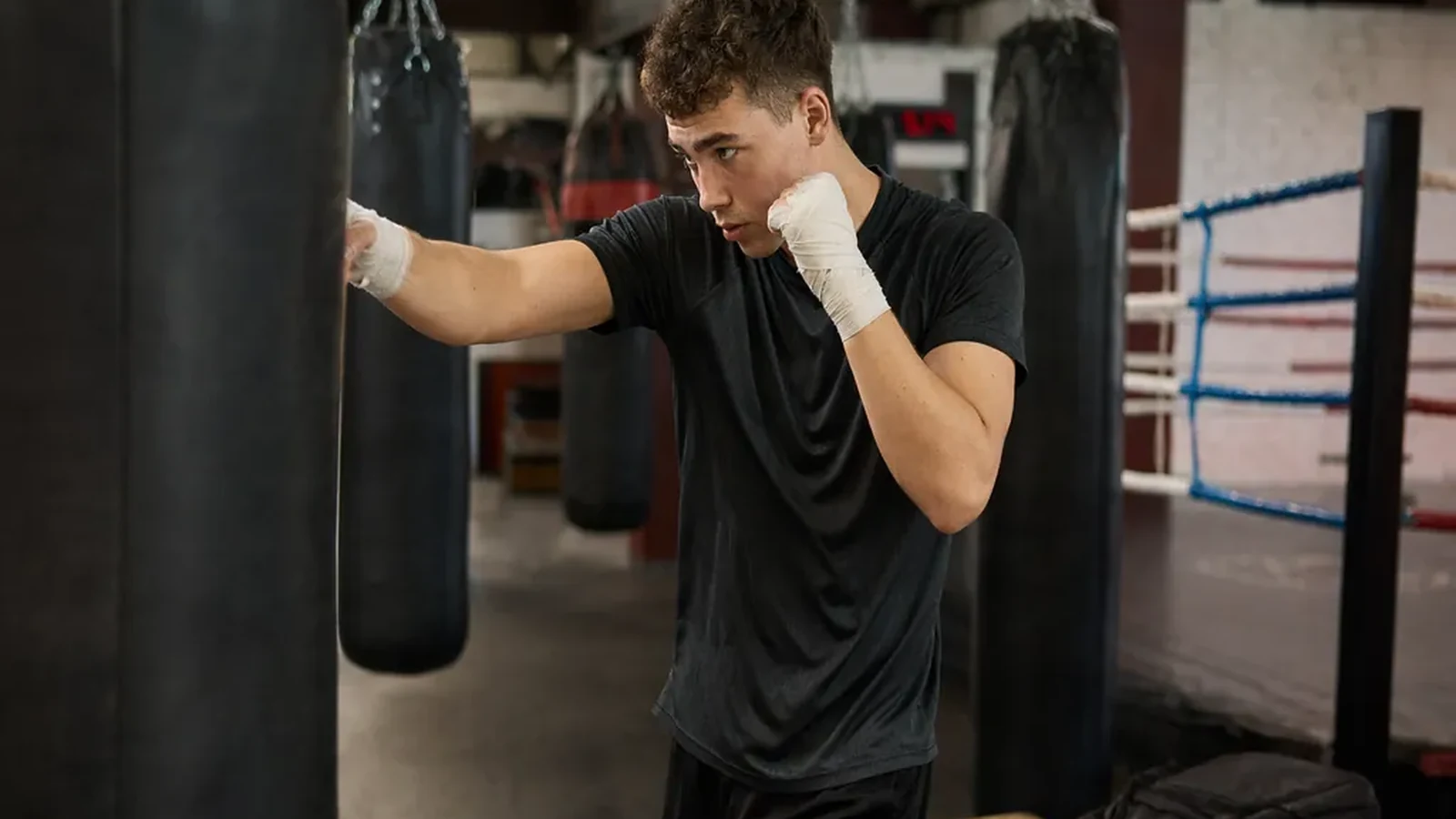Student in dark training kit wrapping hands beside heavy bags before a boxing session