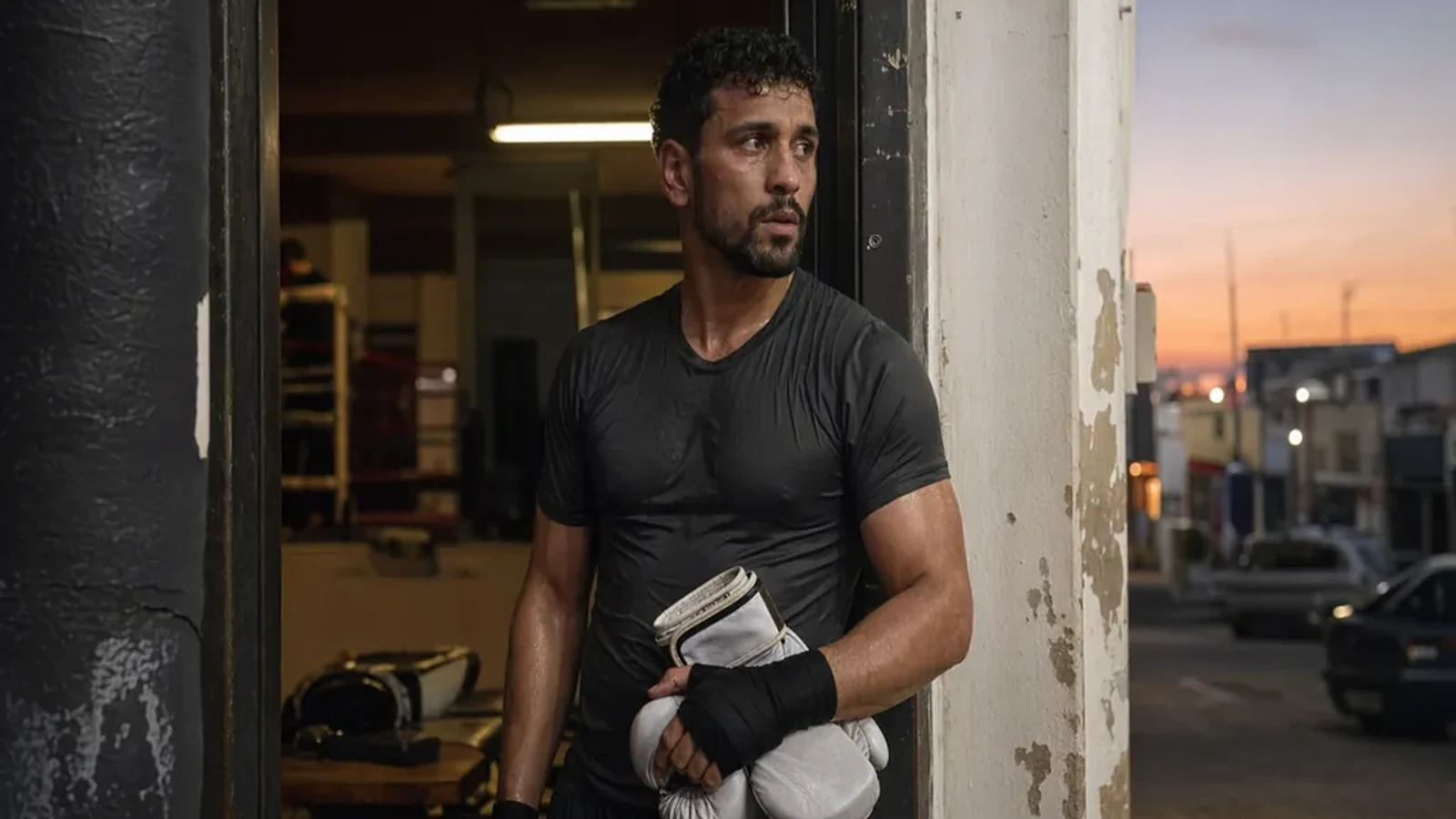 Boxer standing in a gym doorway after training holding white gloves at dusk