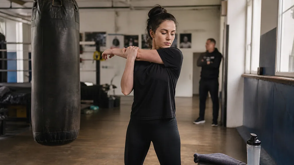 Adult woman doing gentle shoulder mobility beside a heavy bag before boxing training