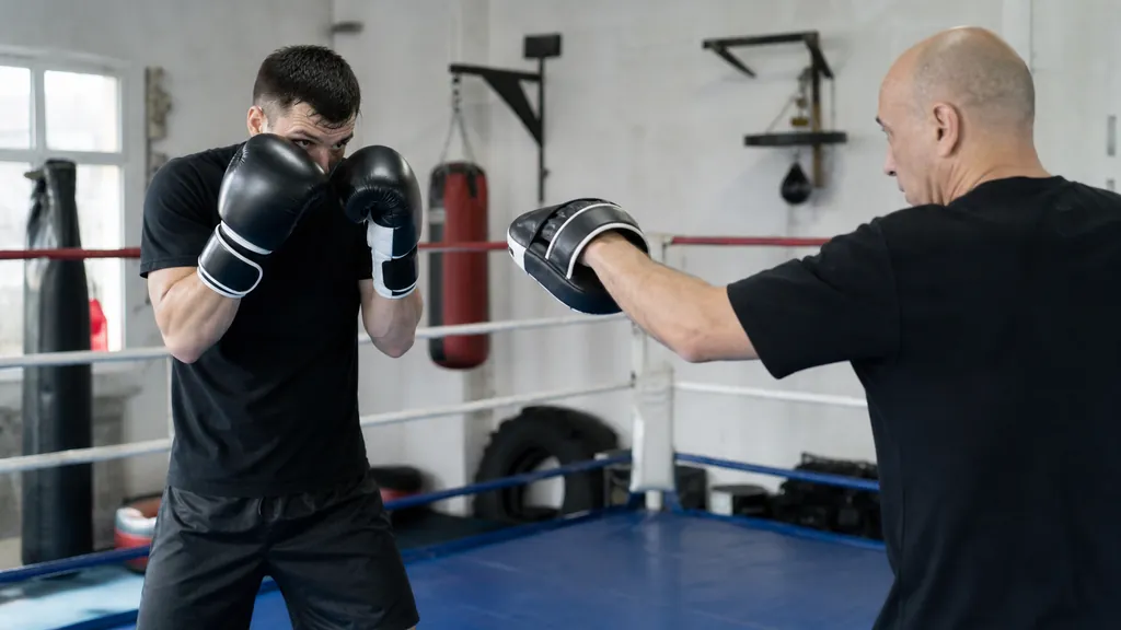 Adult boxer in a compact guard near ring ropes as a coach gives a slow straight-punch mitt cue in a bright community boxing gym