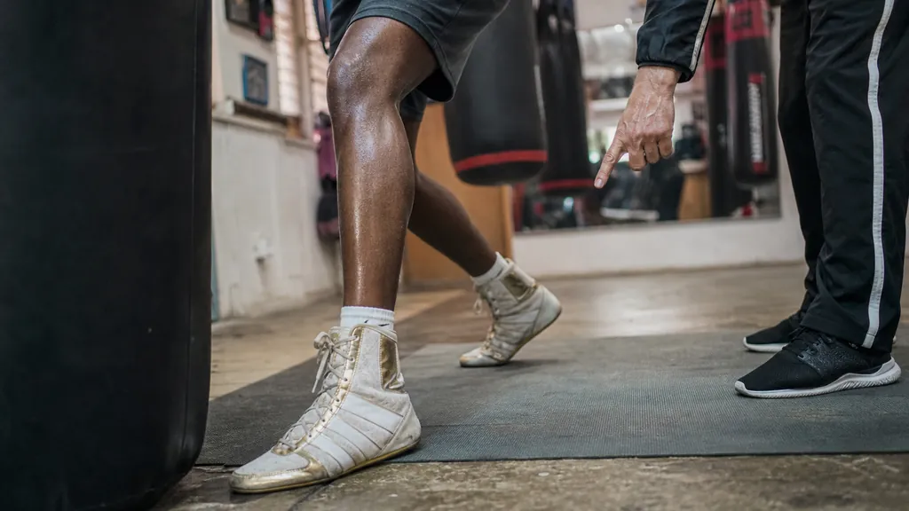 Adult boxer practising a pivot hook on a heavy bag while a coach watches foot placement