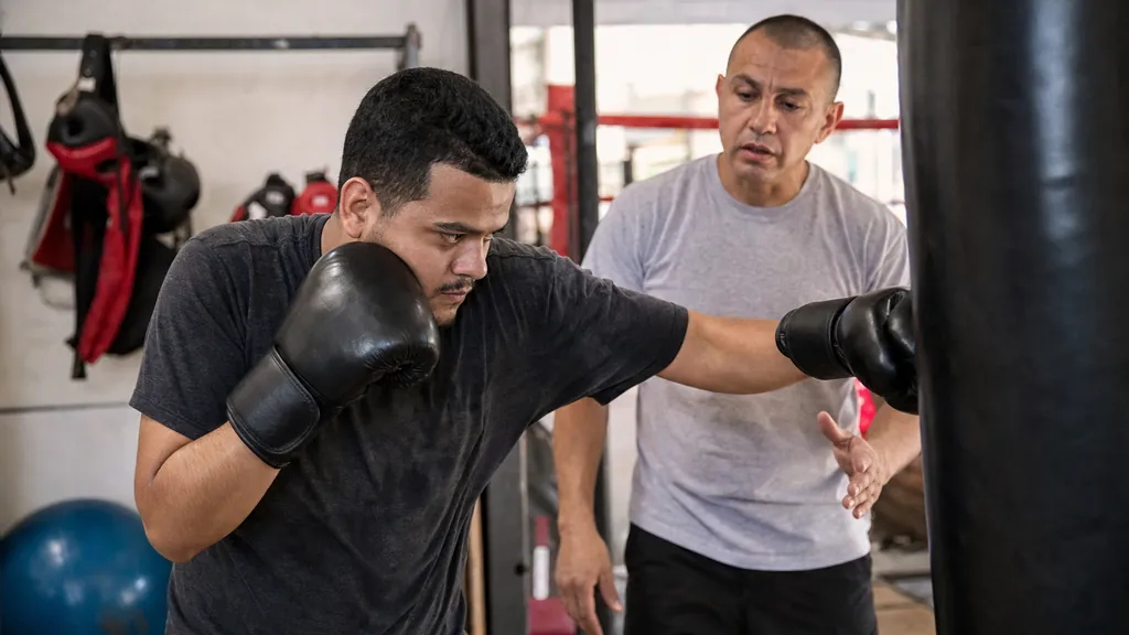 Boxing coach pointing at an adult boxer's foot position beside a heavy bag during a pivot drill