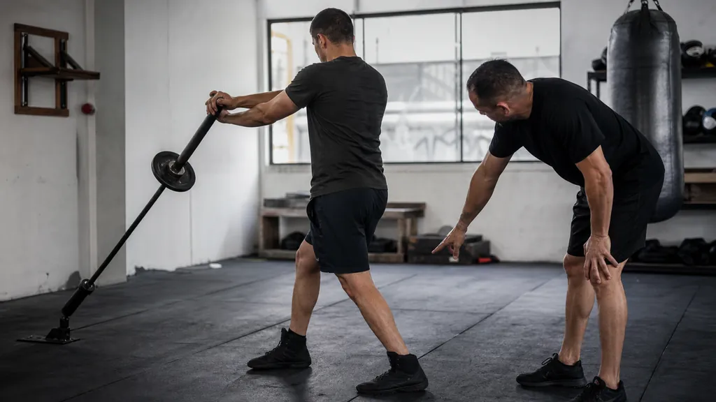 Anonymous adult boxer performing a controlled two-handed landmine rotation while a coach observes in a community boxing gym