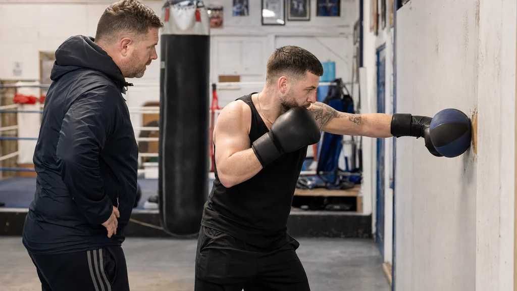 Adult beginner boxer practising a medicine ball wall punch drill under coach supervision