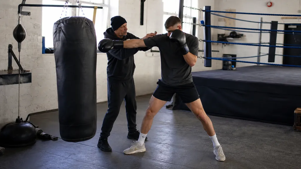 Coach checking an adult boxer's balanced straight-punch position beside a heavy bag during a technical drill