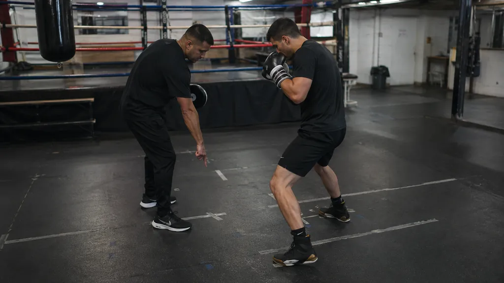 Coach holding focus mitts while an adult boxer completes a small lead-foot step and controlled pivot after a rear straight, both gloves near the face