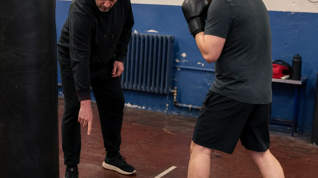 Coach pointing to a floor line beside an adult boxer's staggered stance near a heavy bag