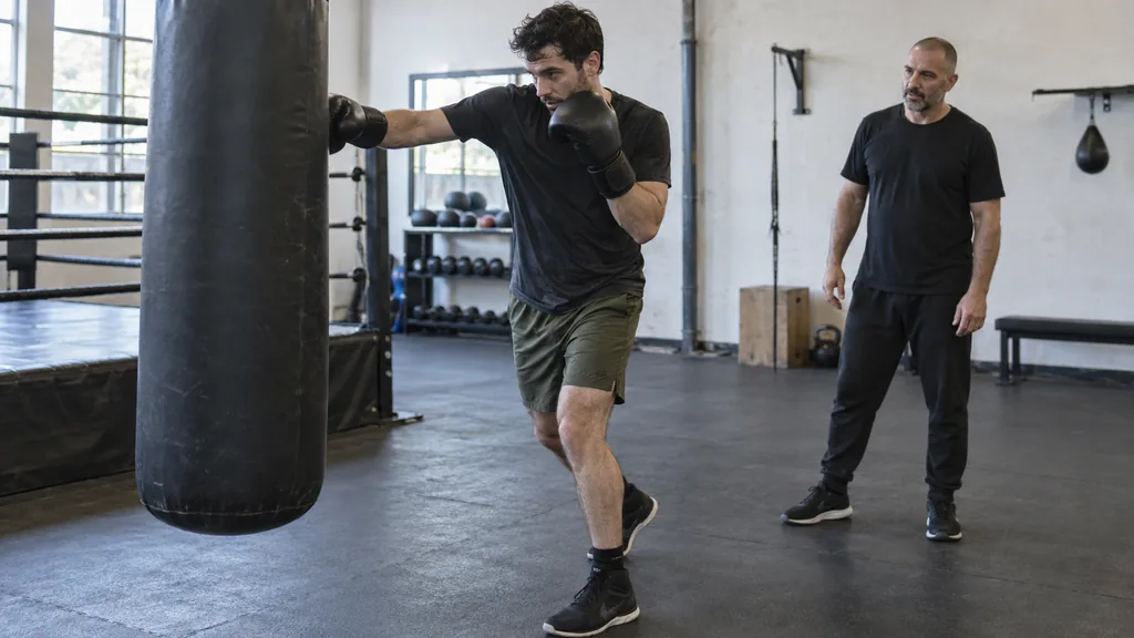 Adult recreational boxer stepping away from a heavy bag after a light jab and returning to a compact balanced guard while a coach observes
