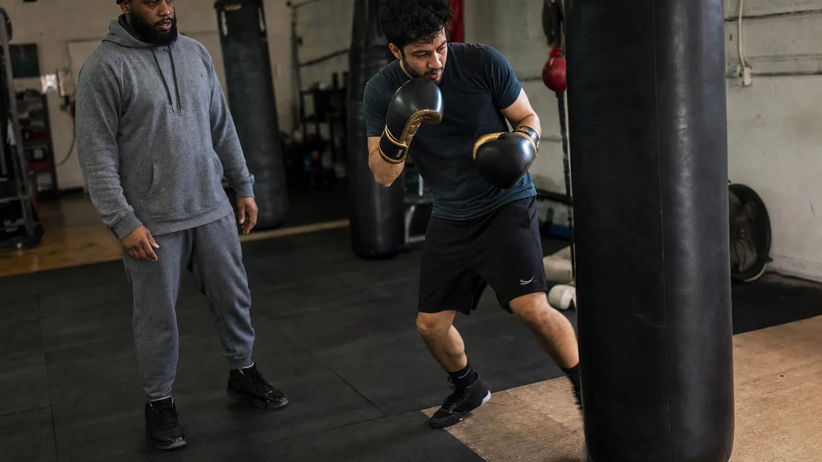 Adult beginner boxer doing purposeful bag work with a coach nearby, focusing on movement after punching