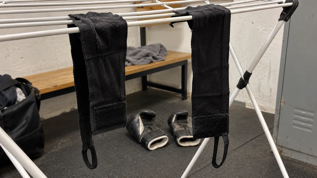 A boxing coach rewrapping a slipped black hand wrap for an adult beginner beside a heavy bag